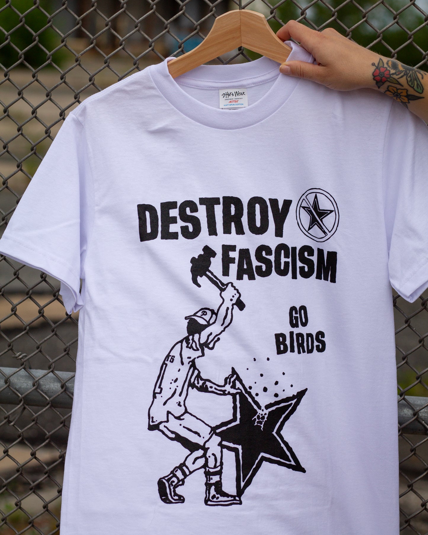 White t-shirt with 'DESTROY FASCISM GO BIRDS' graphic design on a hanger against a chain-link fence background.