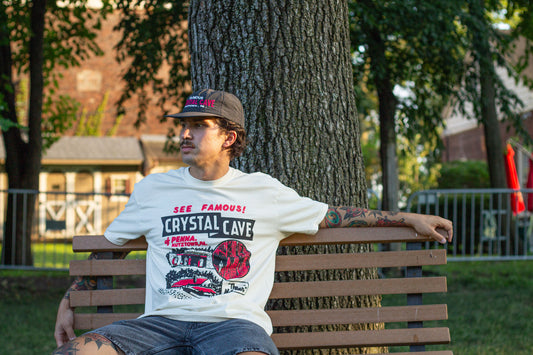 Person sitting on a bench wearing a white t-shirt with graphics and text, in front of a tree.