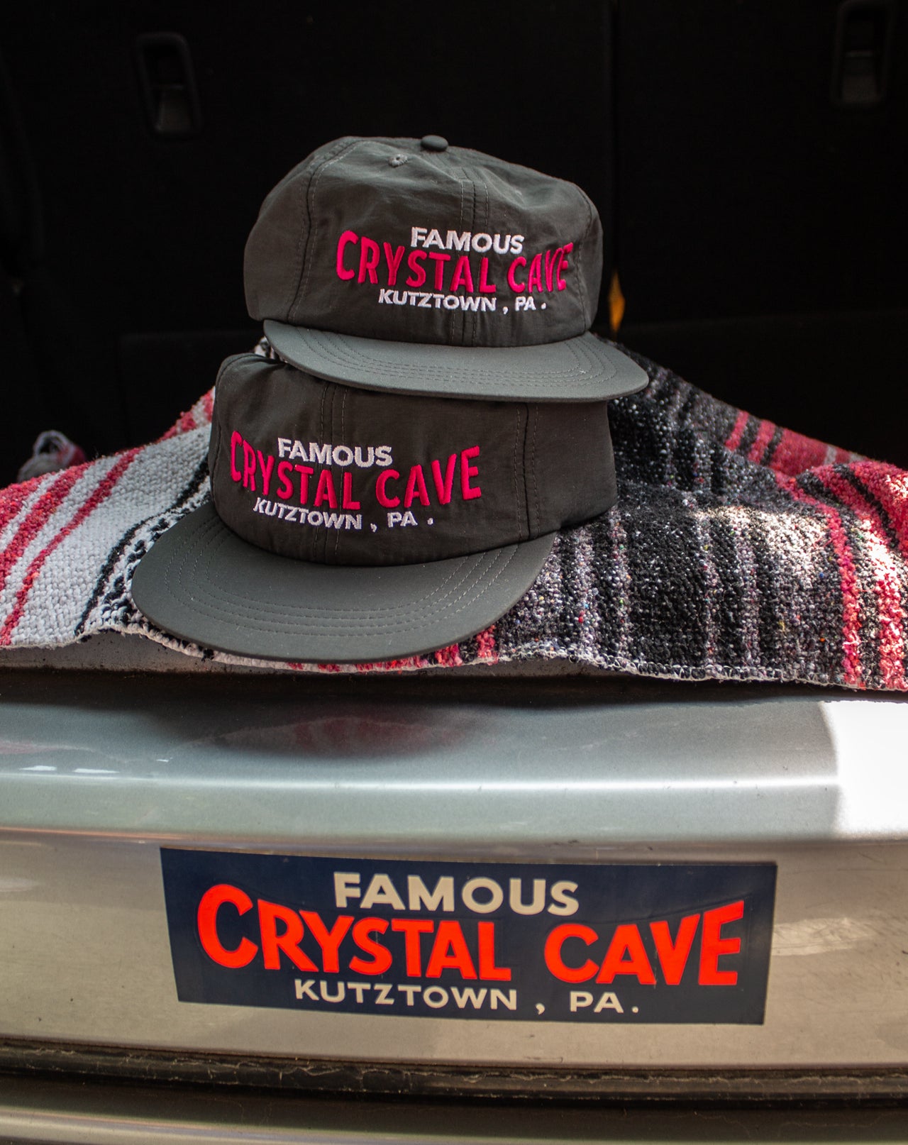 Two black hats with 'Famous Crystal Cave' text on a textured surface with a car bumper in the background.