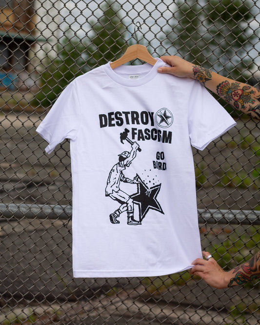 White t-shirt with graphic design and text 'DESTROY FASCISM GO BIRD' held by a person against a chain-link fence background.
