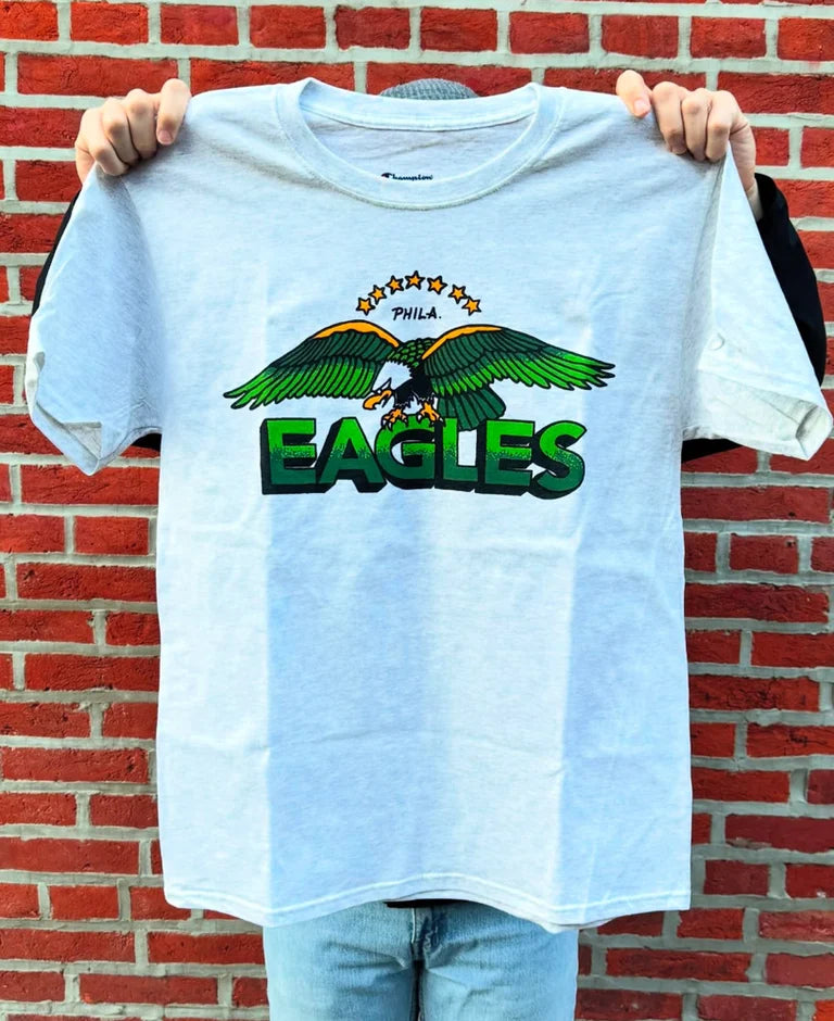 Grey t-shirt with green 'Eagles' logo held up against a brick wall.