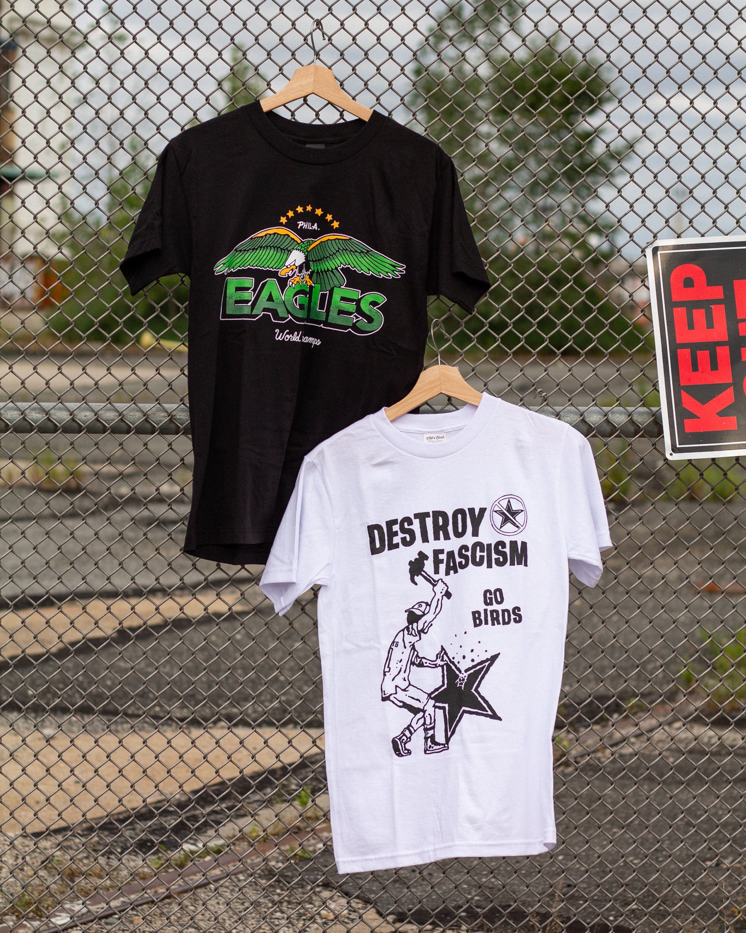 Two t-shirts hanging on wooden hangers against a chain-link fence background.