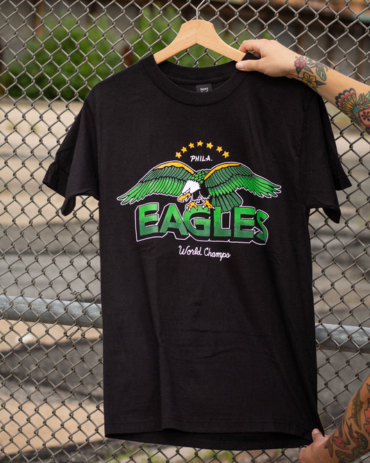 Black t-shirt with green and white eagle graphic and text, held up against a chain-link fence.
