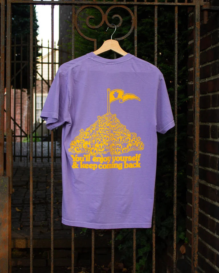 Purple t-shirt with yellow graphic and text on a hanger against a metal gate background