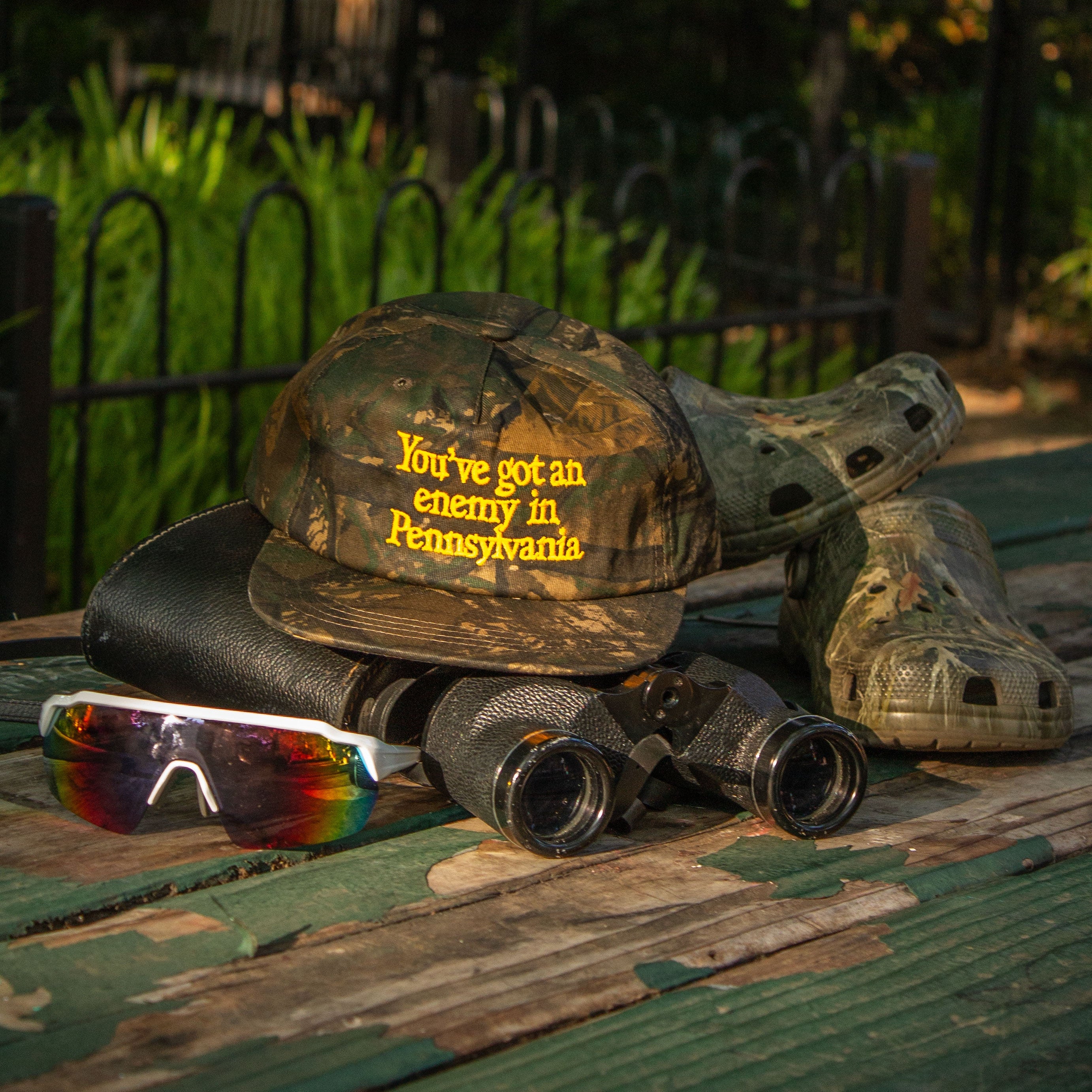 A camouflage baseball cap with yellow embroidered text reading 'You've got an enemy in Pennsylvania' is displayed outdoors on a weathered green wooden bench, alongside a pair of binoculars, sunglasses, and a camouflage boot.