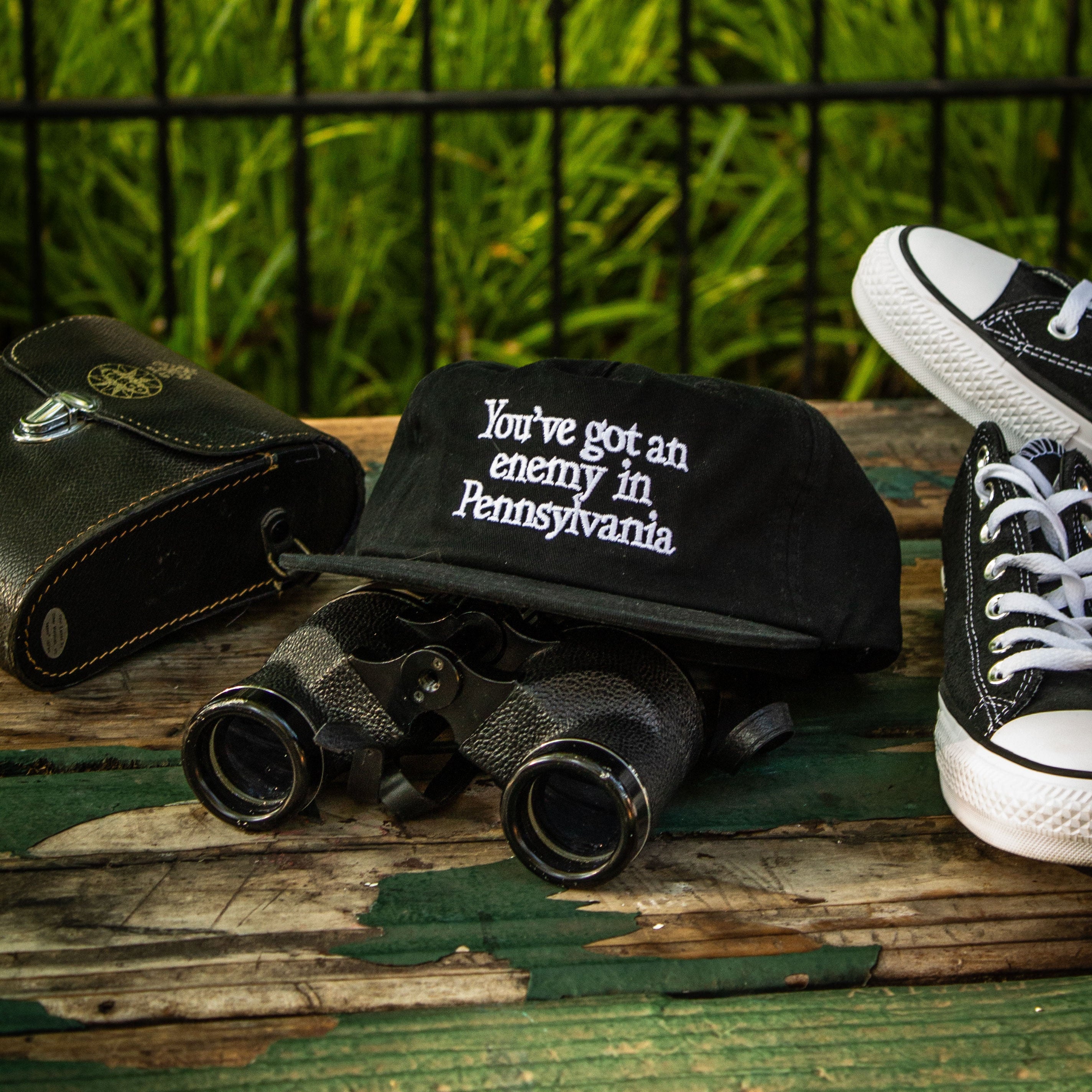 A black fabric cap with white embroidered text reading 'You've got an enemy in Pennsylvania' is displayed on a weathered wooden surface, alongside black binoculars, a black case, and black-and-white sneakers, with a green garden and black fence in the background.