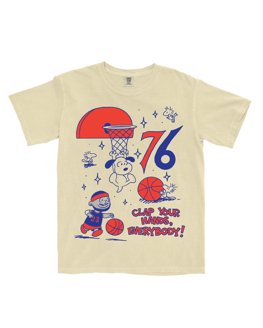 A cream-colored short-sleeve t-shirt featuring a colorful graphic print of Snoopy and Charlie Brown playing basketball, with the text 'CLAP YOUR HANDS, EVERYBODY!' and the numbers '76'.
